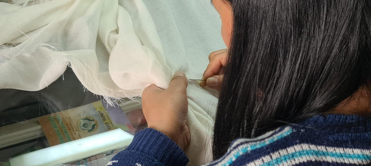 Eine Näherin arbeitet mit der Nadel am Saum eines weißen Kaschmirtuchs; in der Vitrine dahinter ein Chyangra-Pashmina-Etikett